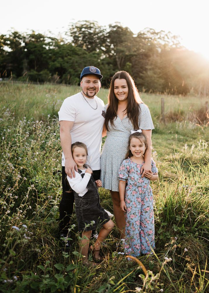 A family poses together in a sunlit meadow during golden hour wearing coordinated casual outfits.
