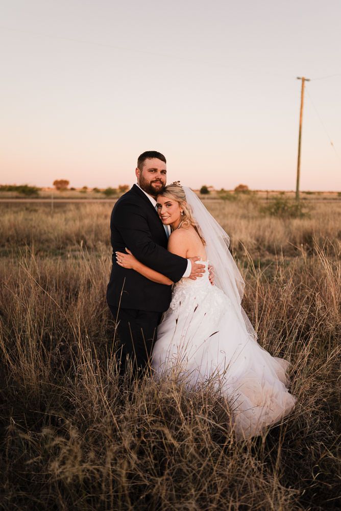 A romantic wedding photo in a rustic field at sunset with tall grass and a utility pole in the background.