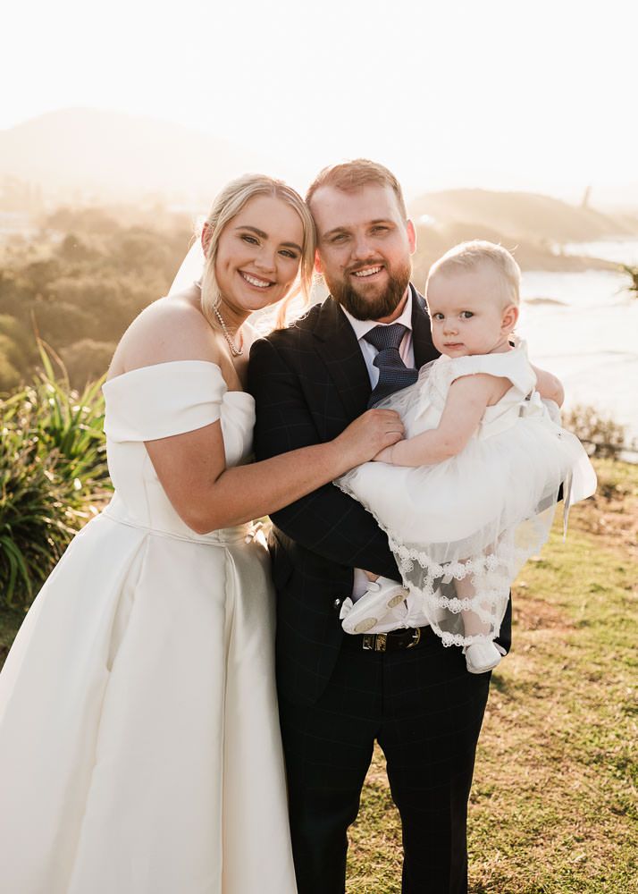 A couple in formal wedding attire embraces outdoors during golden hour on a grassy hilltop with a sunset backdrop.