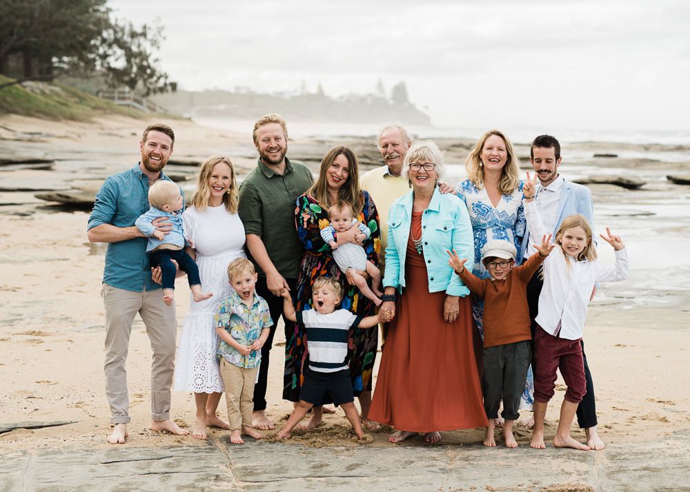 A group of people in coordinated blue and rust-colored outfits pose together on a sandy beach at sunset.