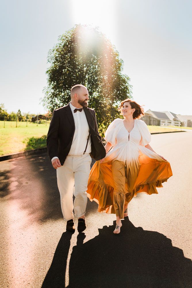 Couple in formal attire dances playfully on a sunlit road, with her orange dress twirling in motion.
