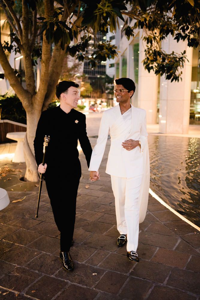 Two people in contrasting black and white outfits walk and interact along a stone pathway under trees at dusk.