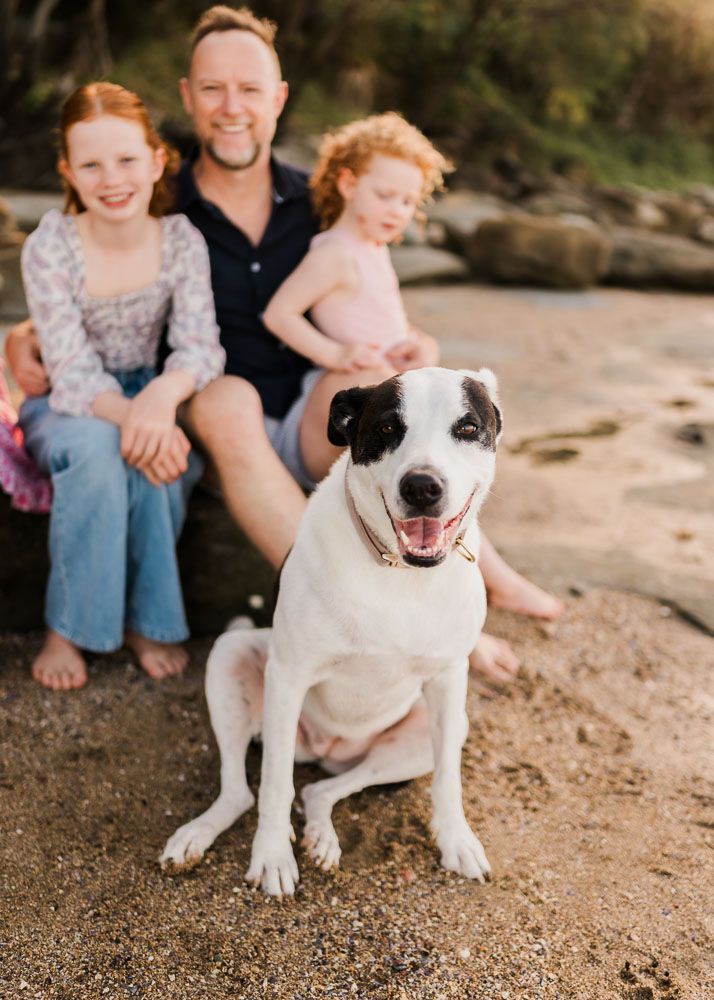 A happy white and black dog sits on a sandy beach with a man and two red-haired girls smiling behind it.