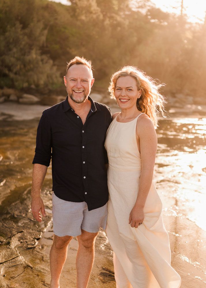 A smiling couple poses together on rocky shoreline at golden sunset, man in black shirt and woman in white dress.