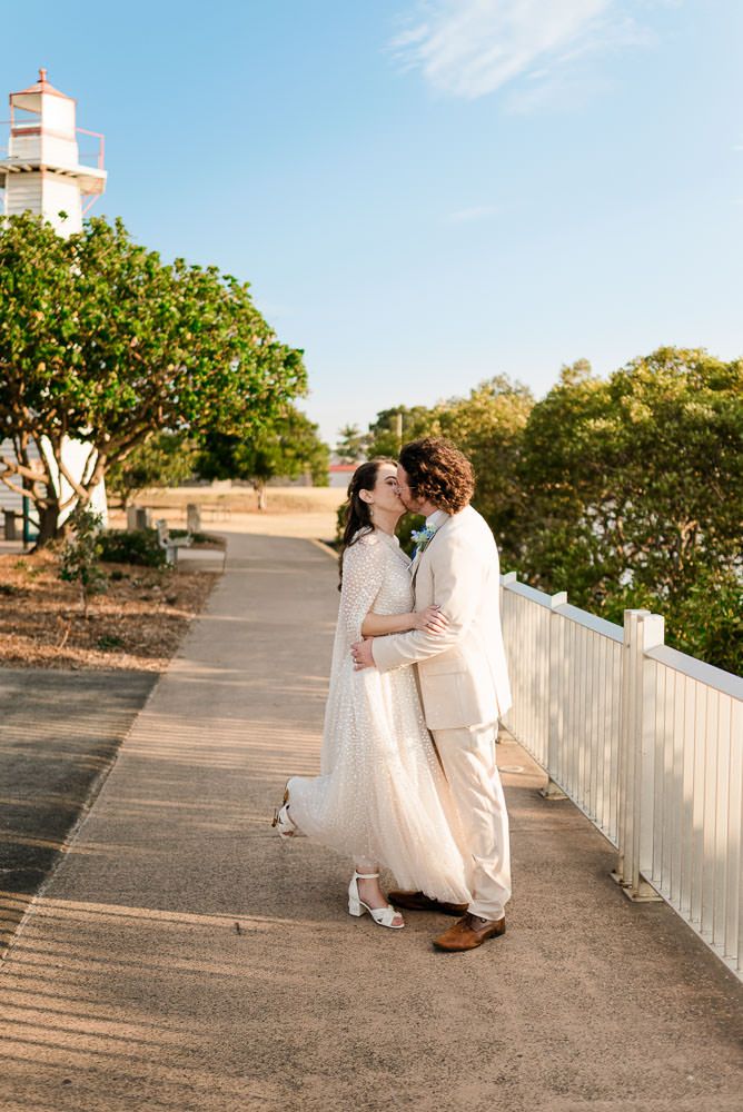 A couple in white attire walks along a pathway with trees and greenery in the background at sunset.