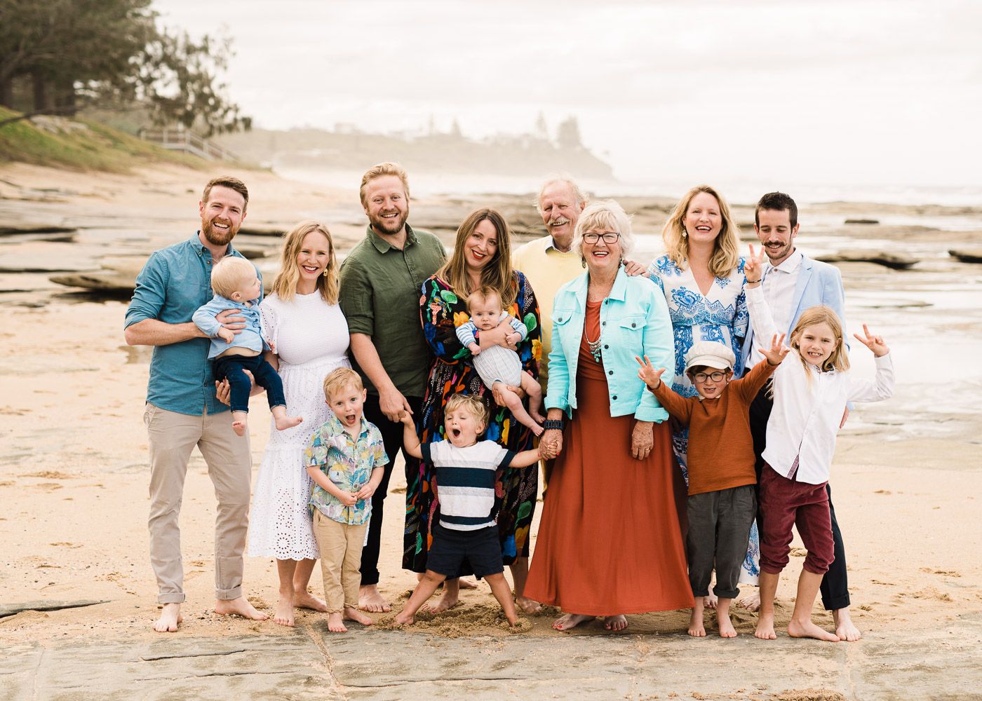 Large family group smiling together on a sandy beach, with ocean and rocky cliffs in the background.