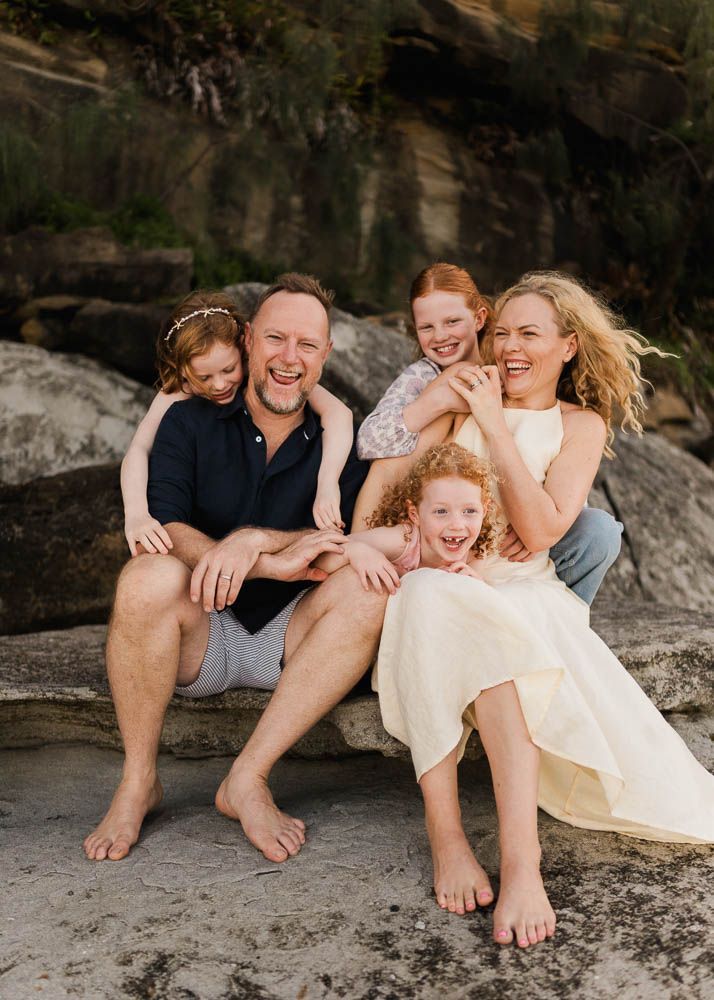 Happy family of five laughing together on rocky beach, parents with three red-haired children enjoying a sunny outdoor portrait.