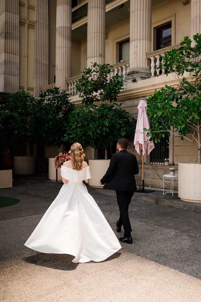 A bride in a white wedding gown and groom in black suit walk together in front of a neoclassical building with columns.