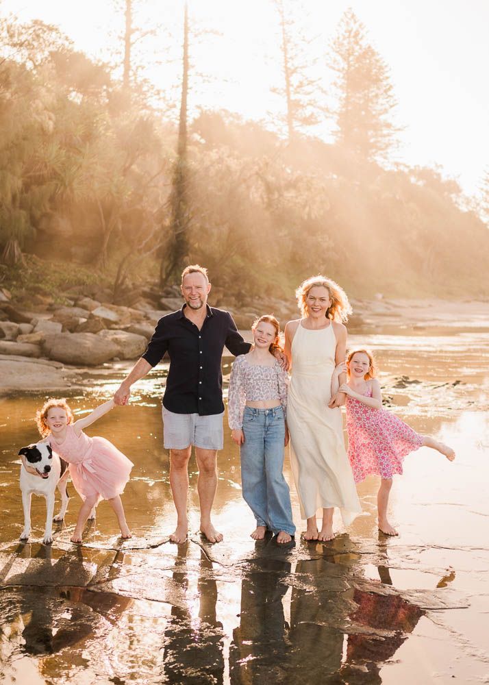 Family of four with a dog walking barefoot on a sunny beach, smiling and enjoying golden hour light.