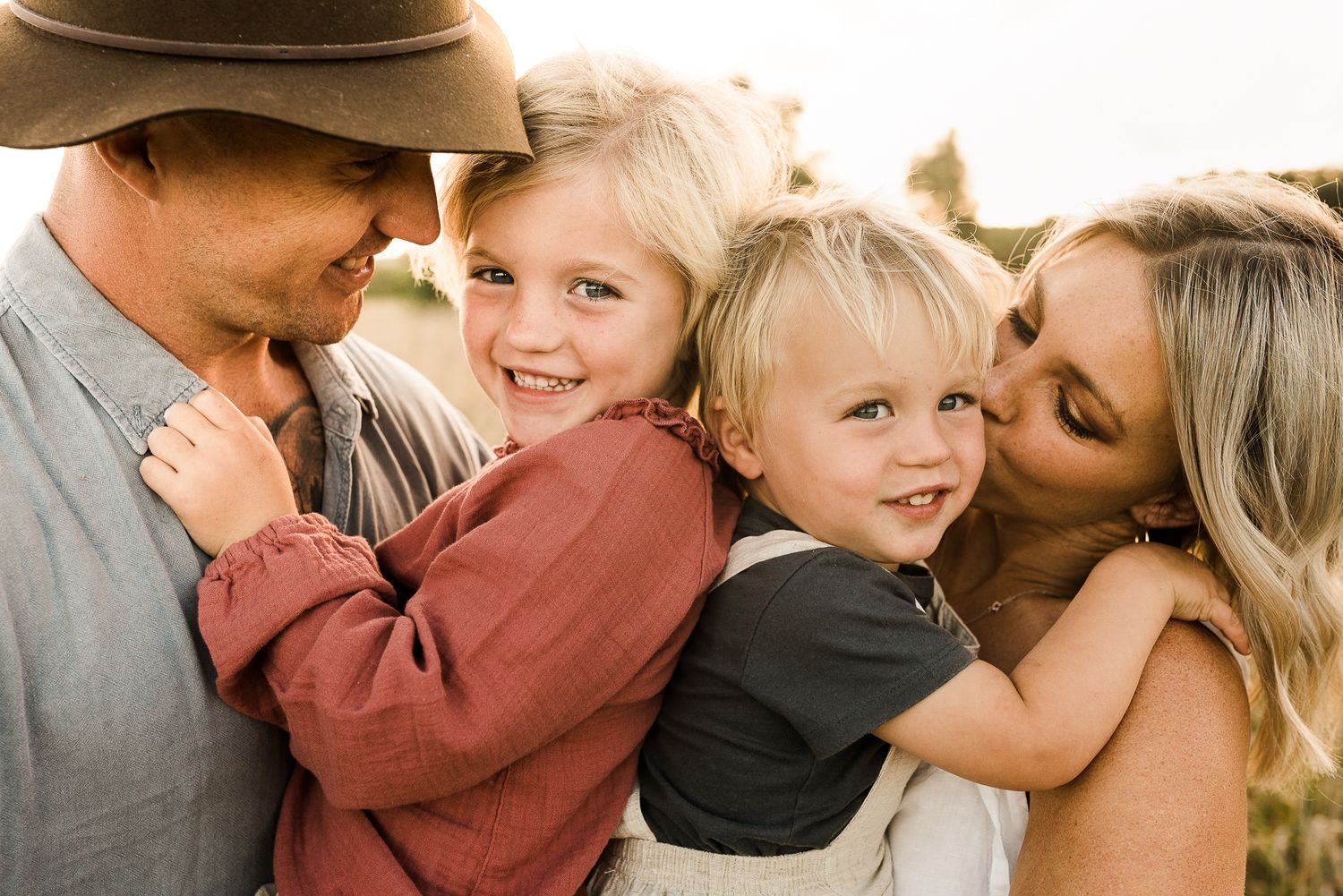 A happy family of four with two young blonde children smiling outdoors in warm golden light.