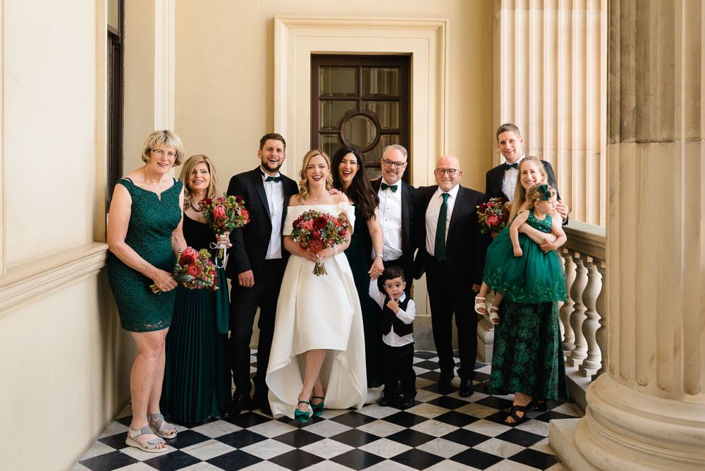 Wedding party members in emerald green dresses and black suits pose along a grand hallway with checkered floors.