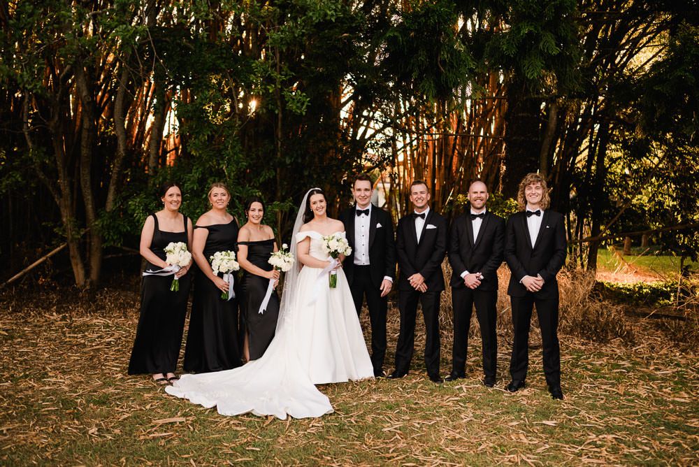 Wedding party poses together in formal attire against a rustic outdoor backdrop with autumn foliage.