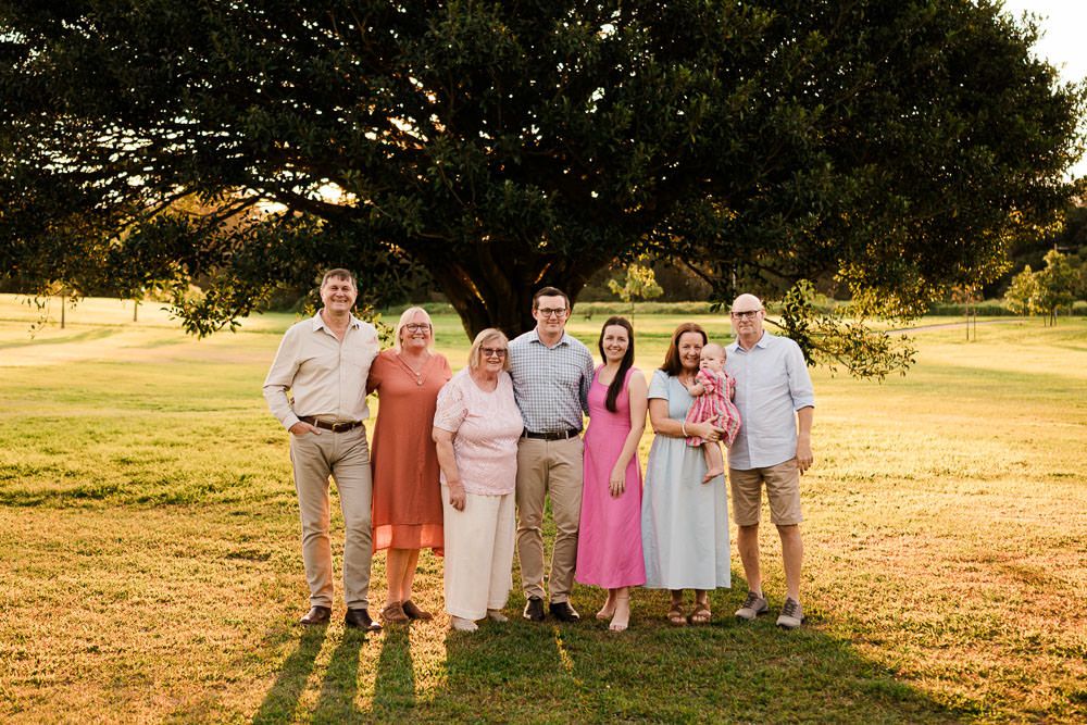 A group in coordinated pastel outfits poses together on a grassy field at sunset under a large tree.