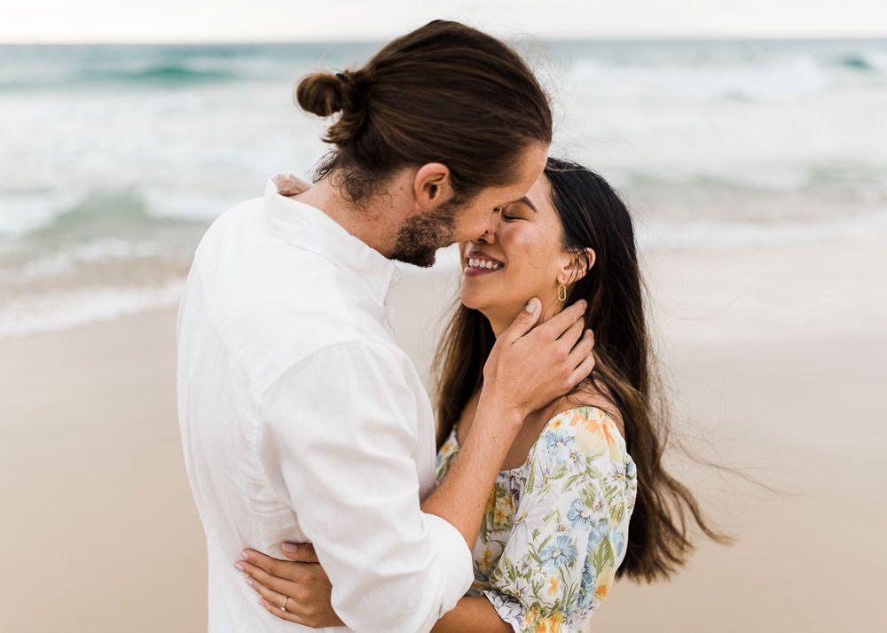 Romantic couple embraces on a sandy beach with ocean waves in the background during sunset.