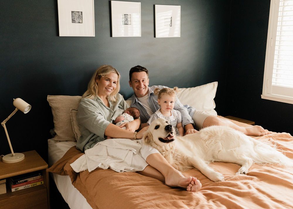Family relaxing together on bed with dog in cozy bedroom with dark walls and pink bedding.