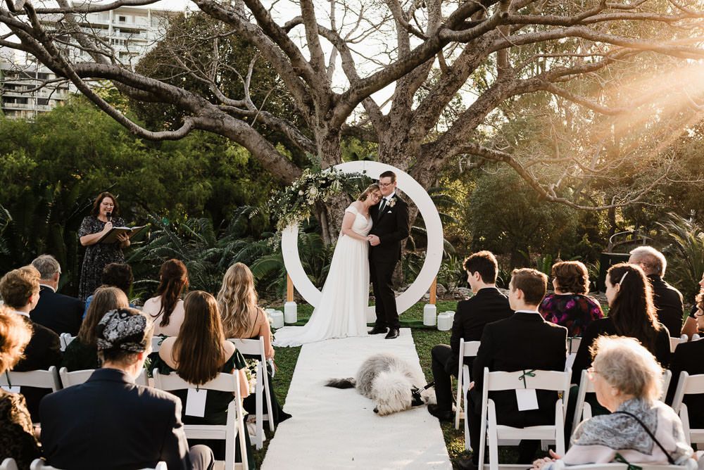 A wedding ceremony takes place outdoors at sunset with guests seated in white chairs and trees in the background.
