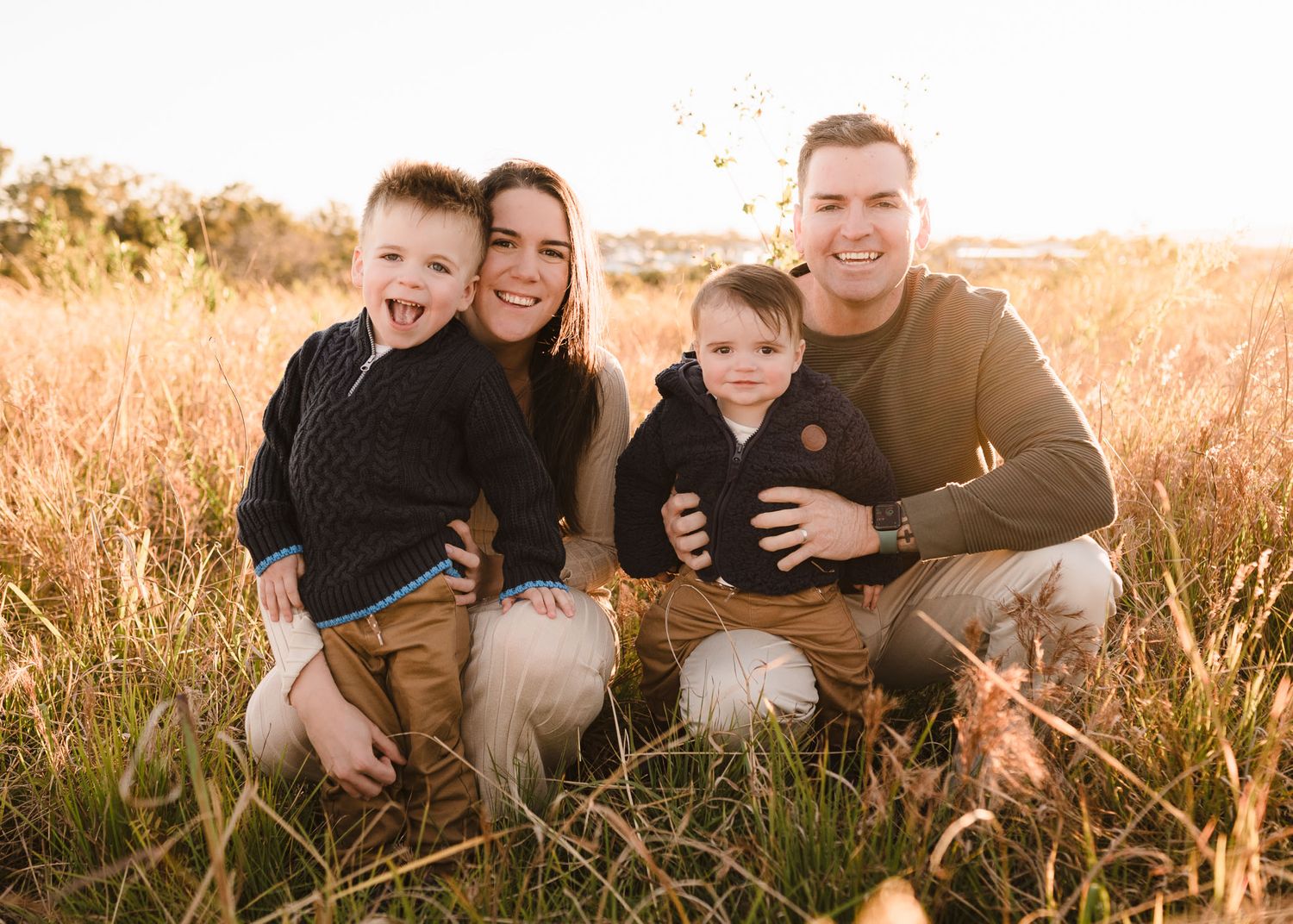 Happy family of four posing in golden field at sunset, parents holding young boys, smiling outdoors.