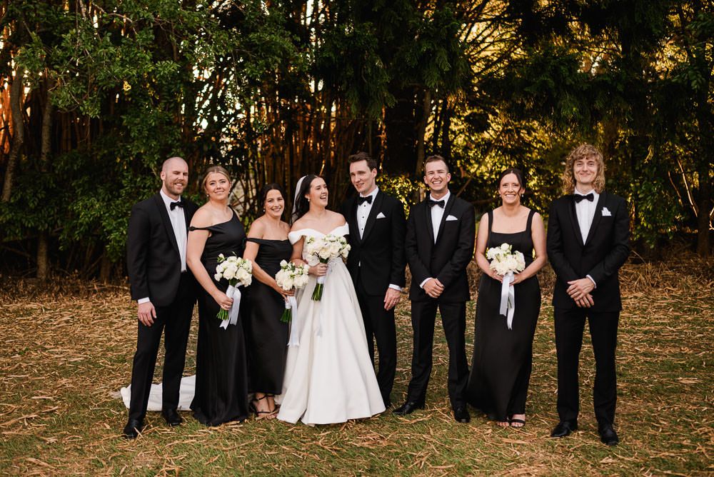 A formal wedding party in black and white attire poses together outdoors in front of autumn trees.
