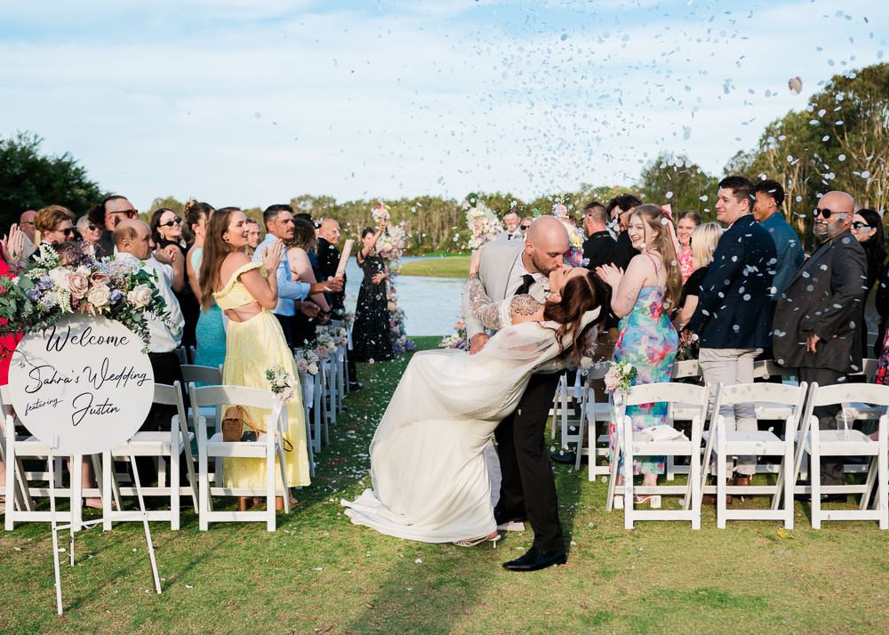 A panoramic view of a wedding ceremony setup outdoors with white chairs and guests gathered on a sunny day.