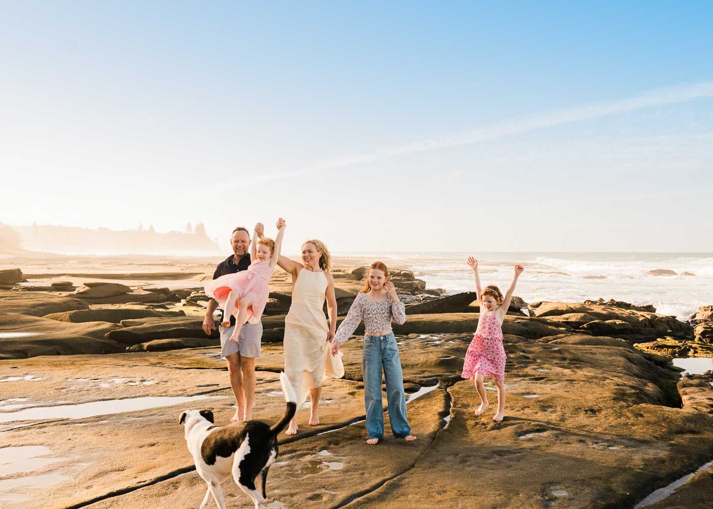 Family with dog celebrating on rocky beach with ocean waves and sunny sky.