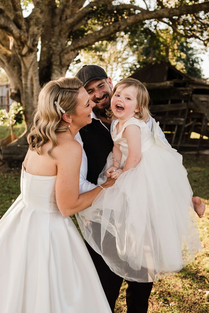 A joyful moment captured outdoors with a young child in a white dress laughing with family during a special occasion.