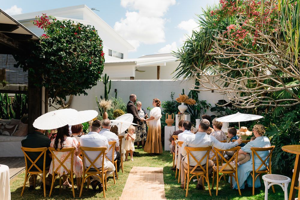 Outdoor wedding reception setup with wooden cross-back chairs and white umbrellas arranged in rows under a sunny sky.