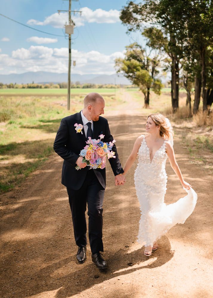A playful couple runs and dances together on a dirt road lined with trees on a sunny afternoon.