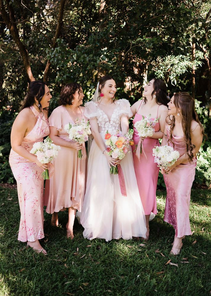 A group of bridesmaids in pink dresses share candid laughs and conversations during a wedding photoshoot outdoors.