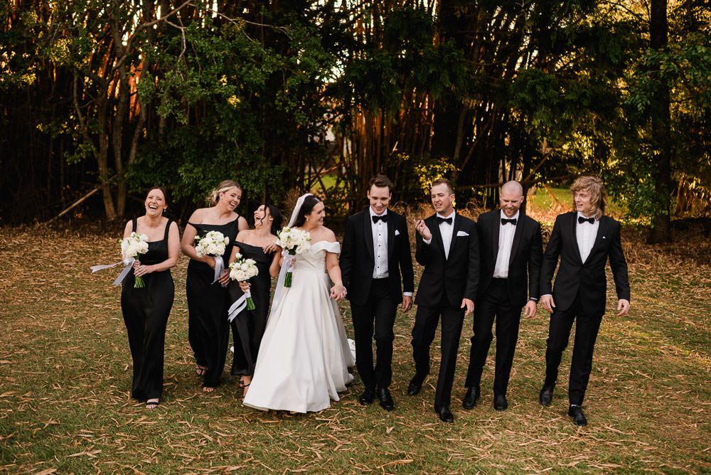 Wedding party members walk and laugh together through a scenic autumn forest setting during golden hour.