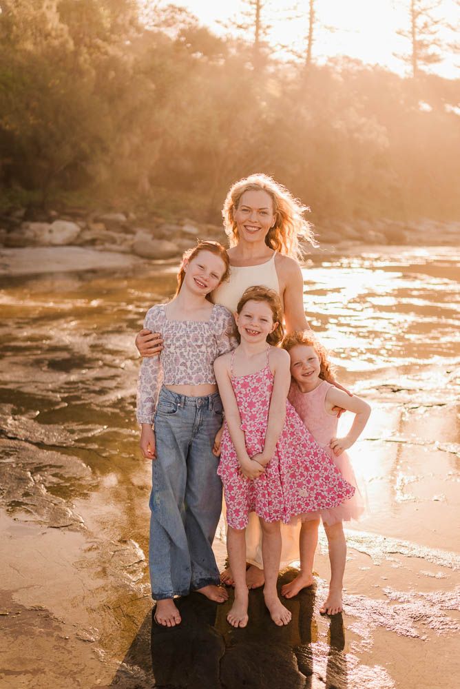 A smiling woman and three young girls pose together on rocks by a sunlit river in pink and casual summer outfits.