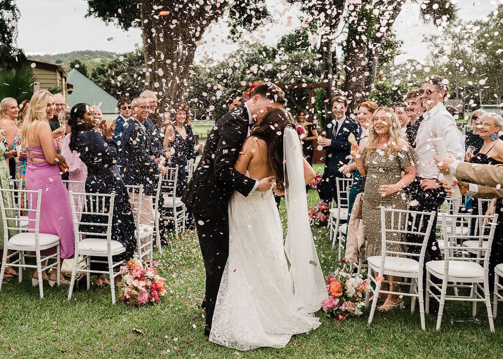 Wedding guests celebrate and dance together outdoors during a joyful reception celebration in a garden setting.