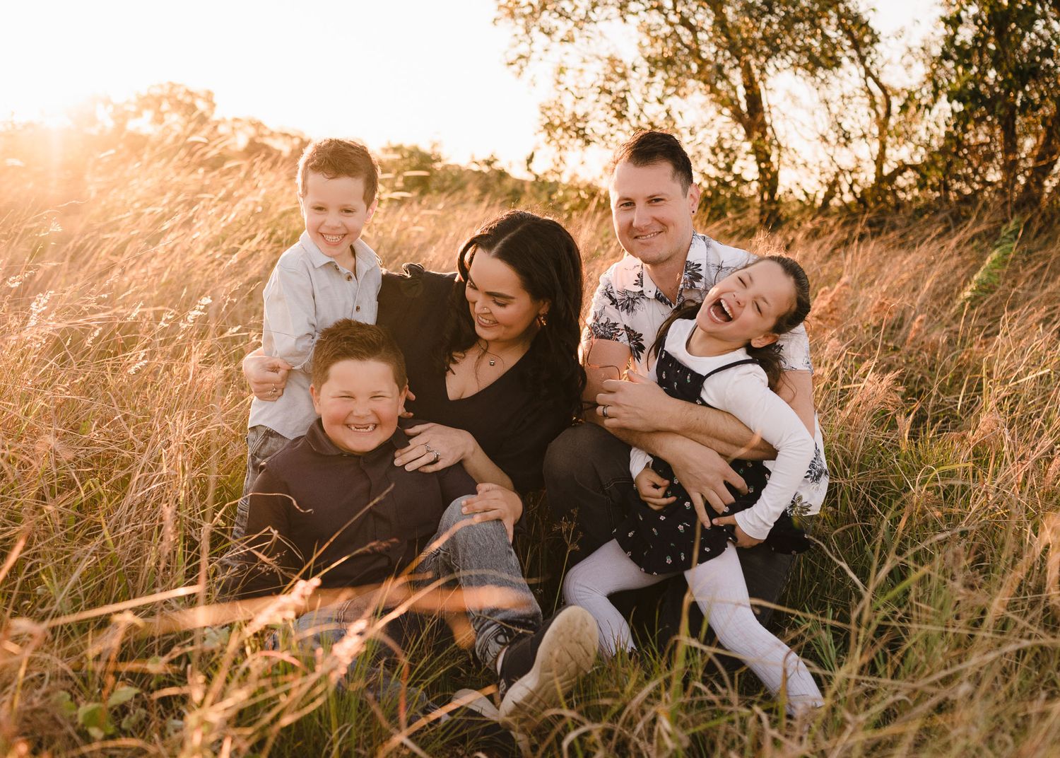 Happy family of five laughing together in golden sunlit field, parents with three children enjoying outdoor portrait session.