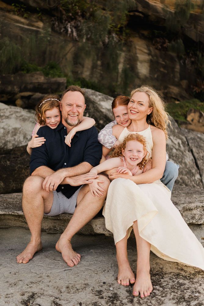 Happy family of five sitting on beach rocks, parents hugging three red-haired children, smiling outdoors near cliff.