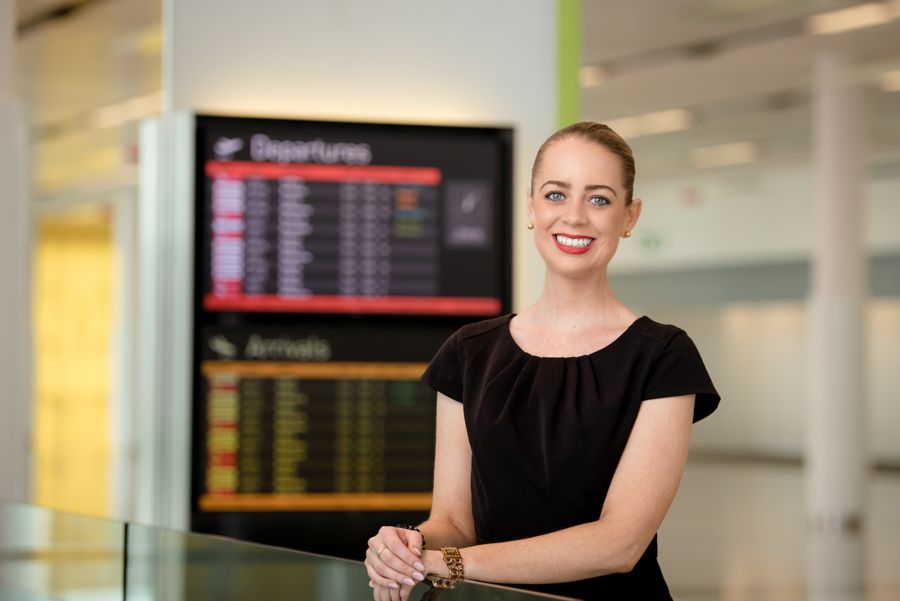 Airport terminal scene with staff member in black attire standing near departure boards.