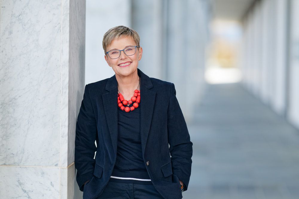 Woman smiling in navy blazer and red beaded necklace leaning against marble column outdoors.