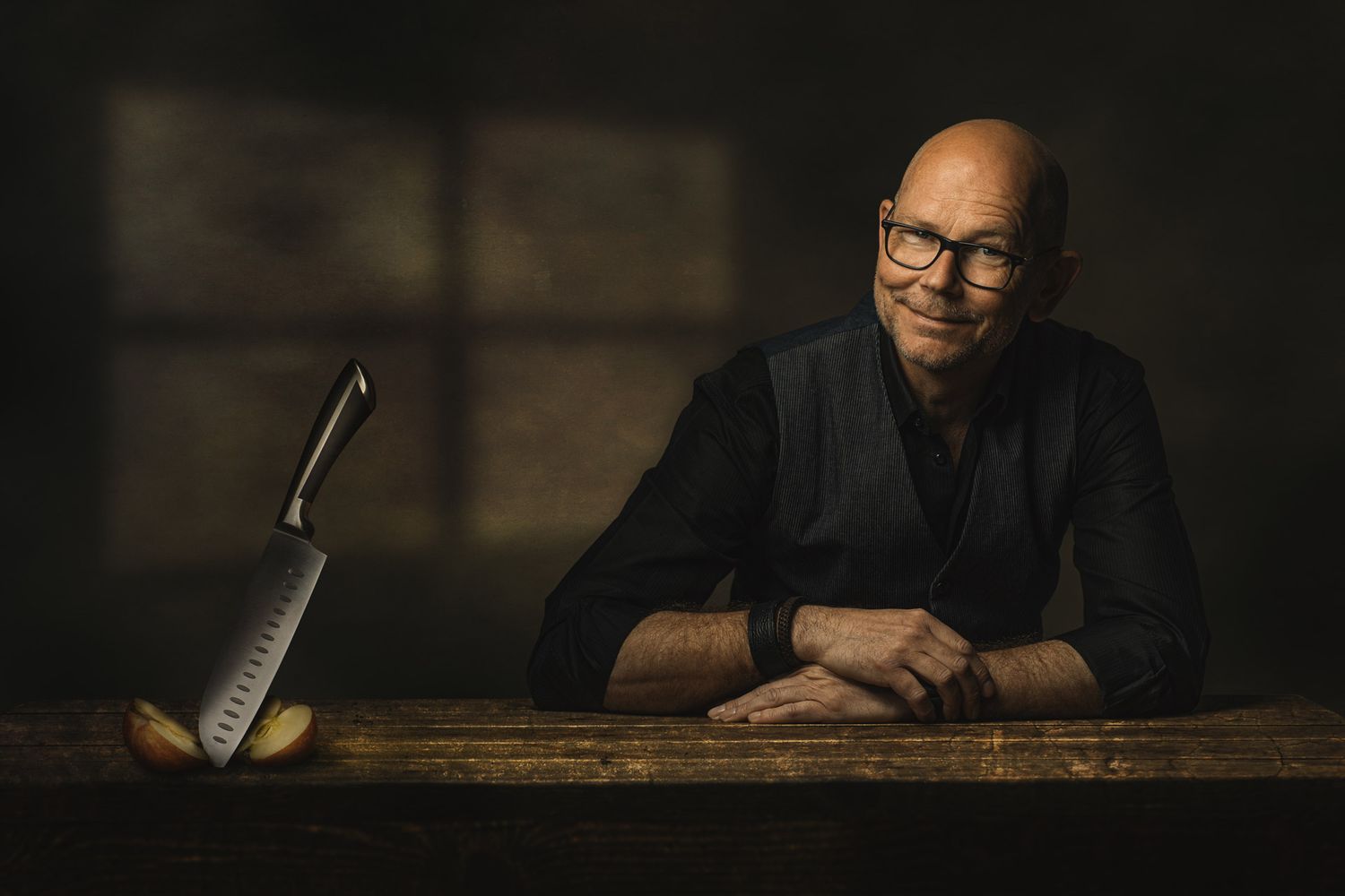Moody portrait of person at wooden desk with knife in dark setting.