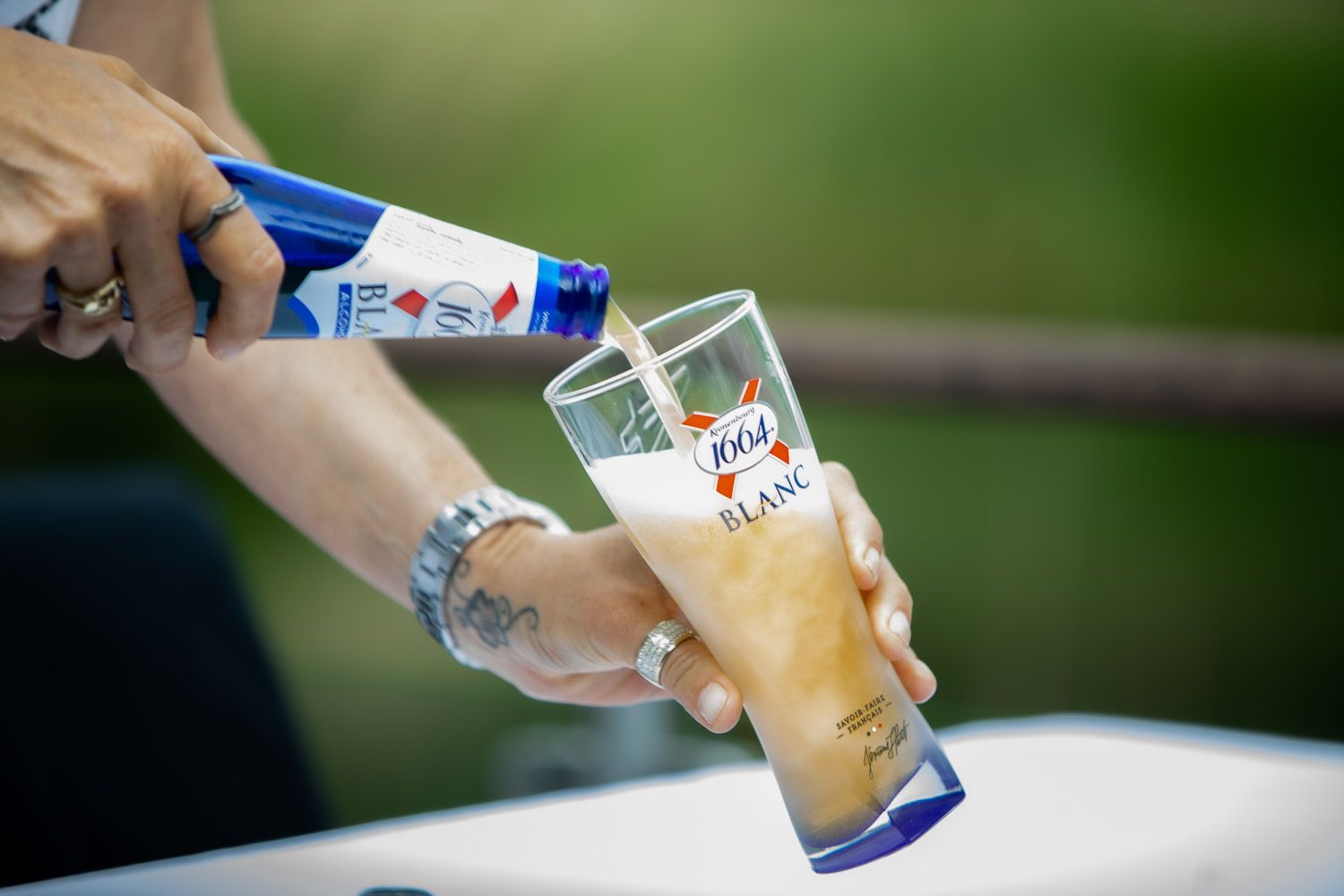 Hands pouring drinks into glasses outdoors with blurred green background.