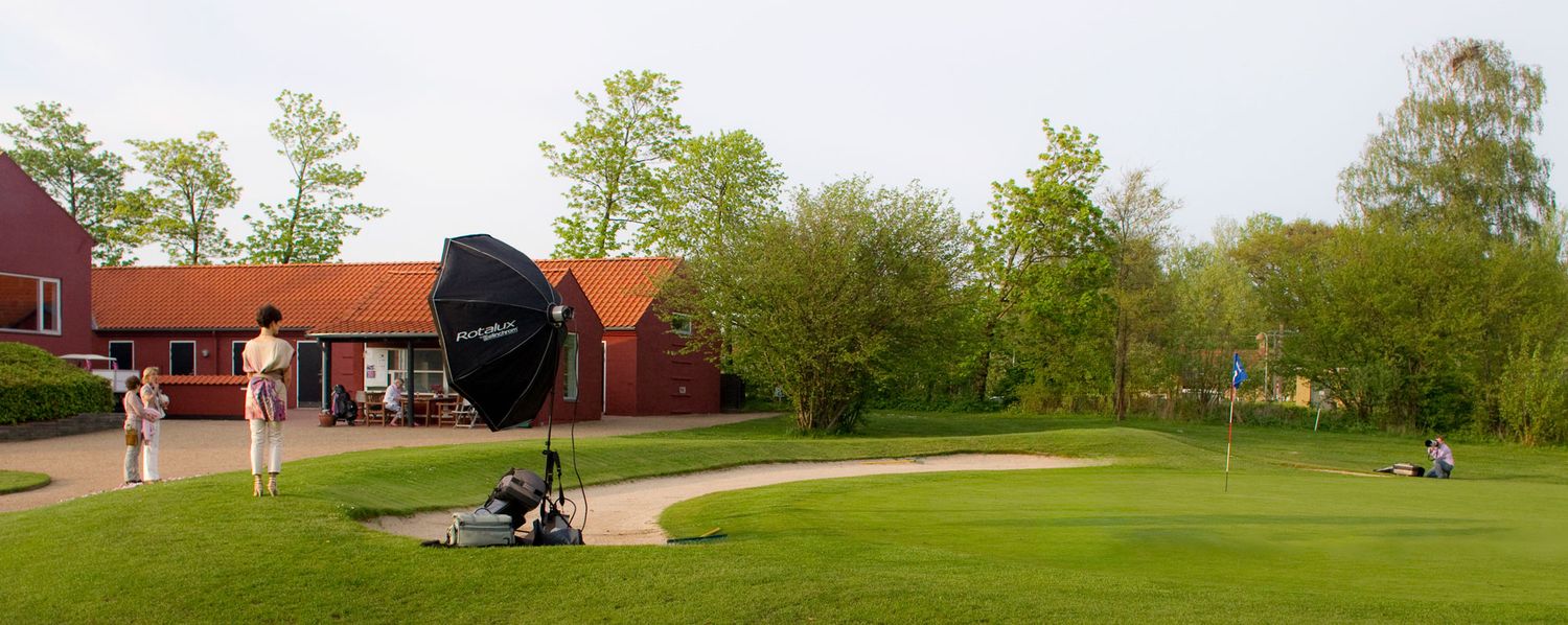 Panoramic view of a golf course with red buildings and trees in the background.