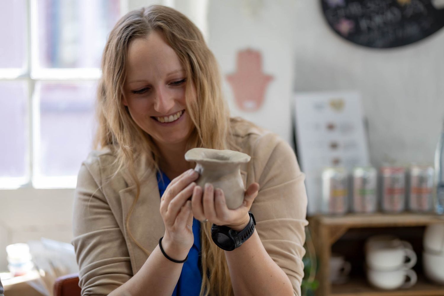 A person in a beige sweater smiles while holding a ceramic mug in a pottery studio.