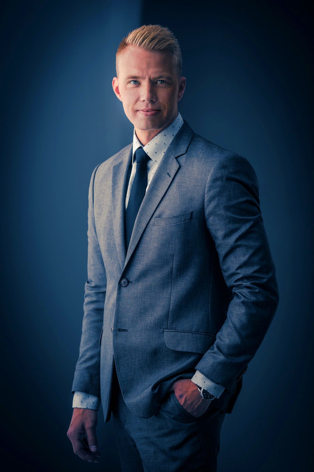 Corporate headshot series in grey suit against deep blue studio backdrop.
