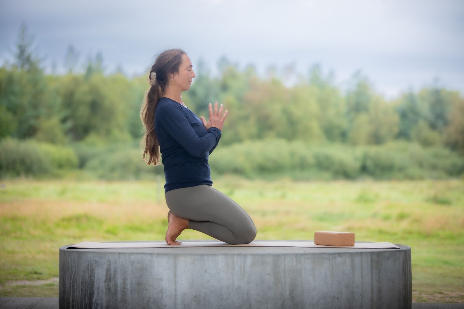 Person sitting in yoga pose on concrete structure outdoors with green trees in background.