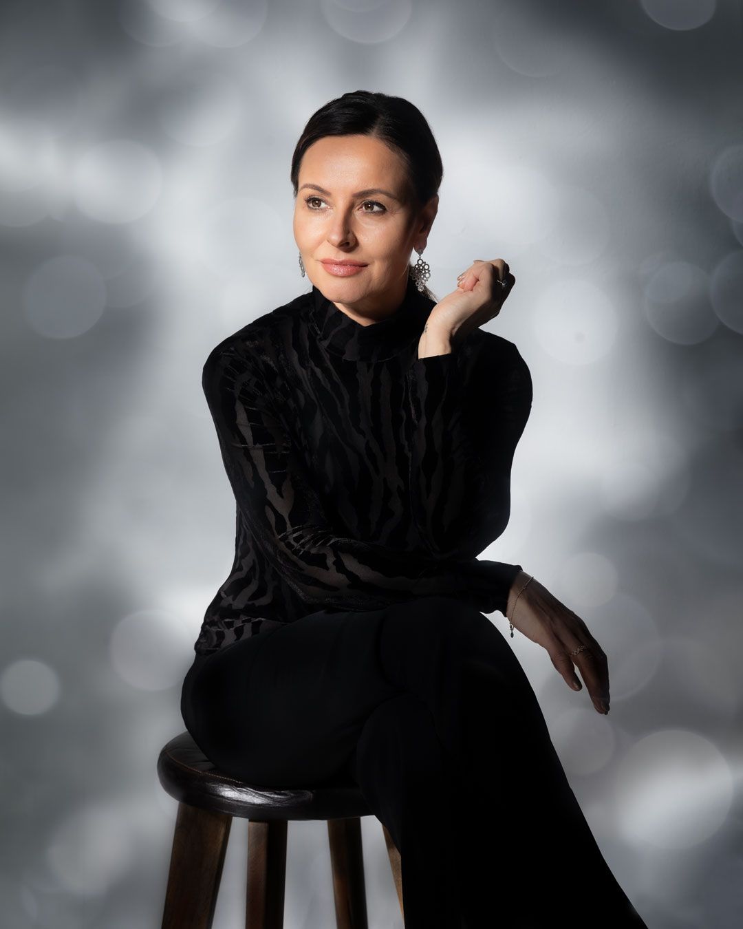 Portrait of woman in black velvet dress seated on stool with bokeh background.