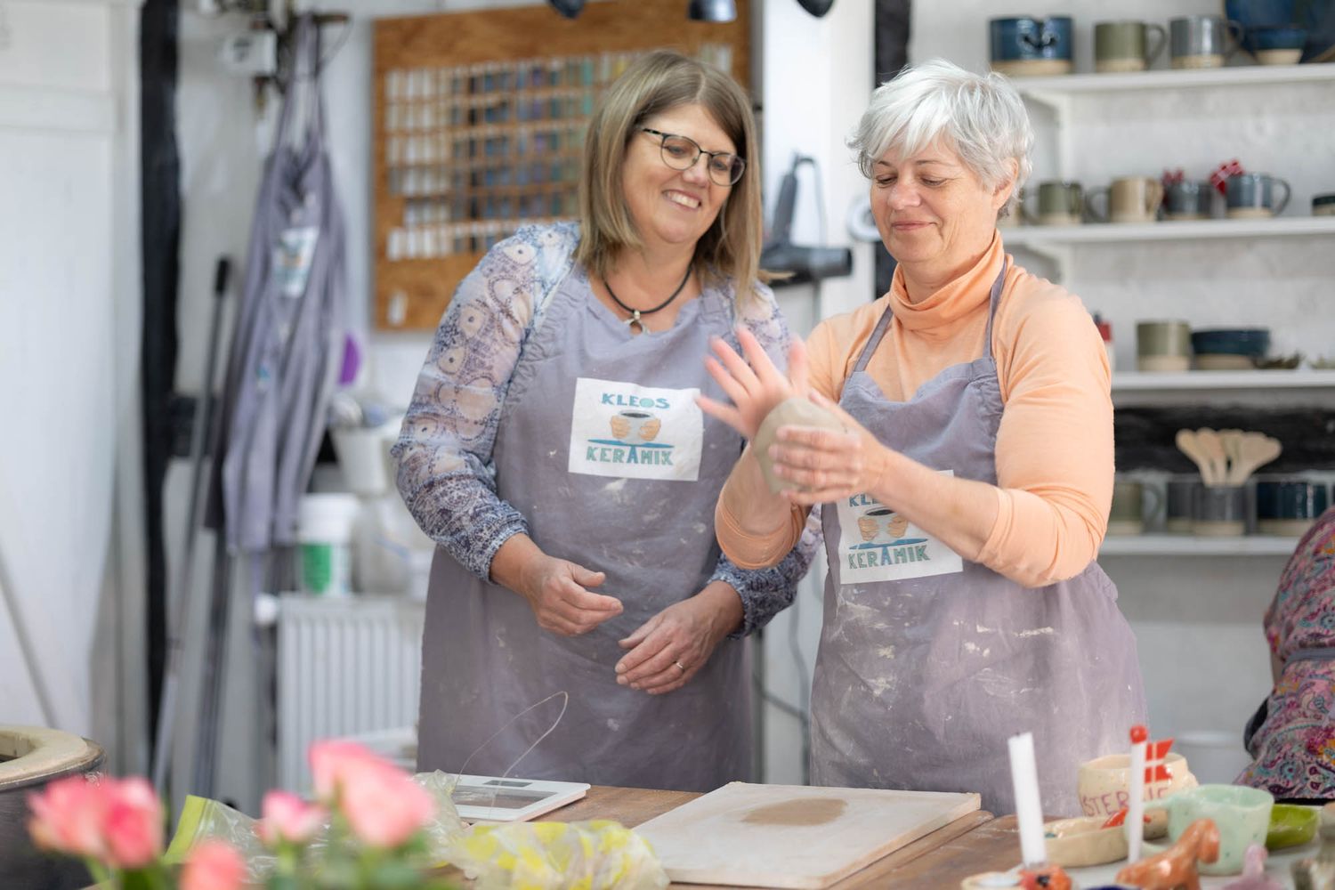 Two people wearing aprons discuss pottery techniques in a bright studio space.