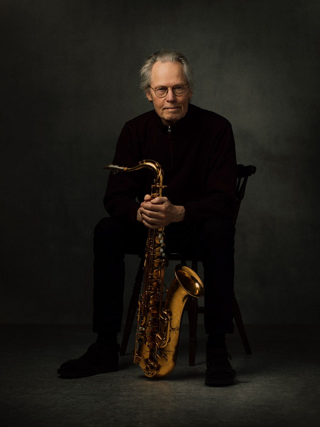 Professional musician seated with saxophone in artistic low-key portrait against dark background.