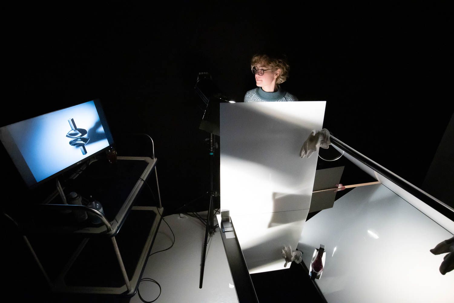 Photographer working with reflective surfaces and studio lighting equipment in dark setting.