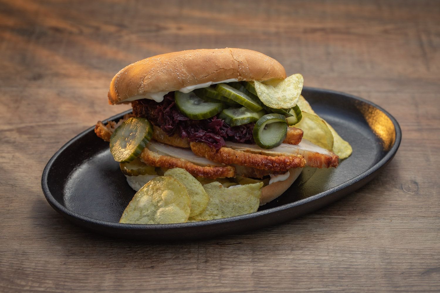 Sandwich with bacon and pickles served on a black plate on wooden surface.