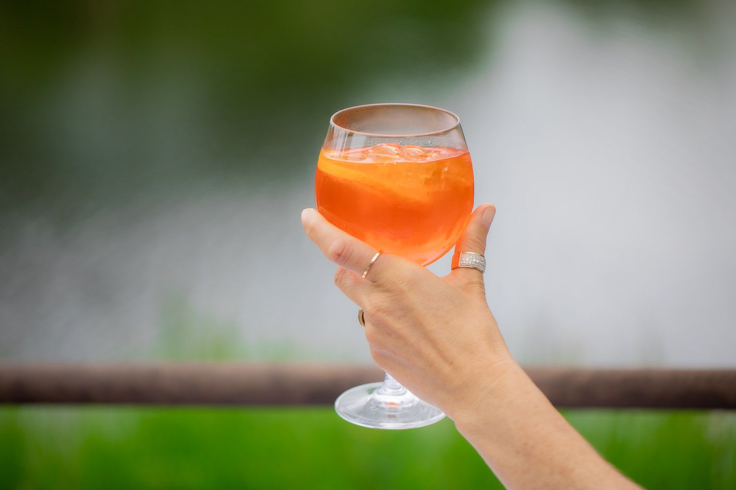 Hand holding orange cocktail against blurred green outdoor background.