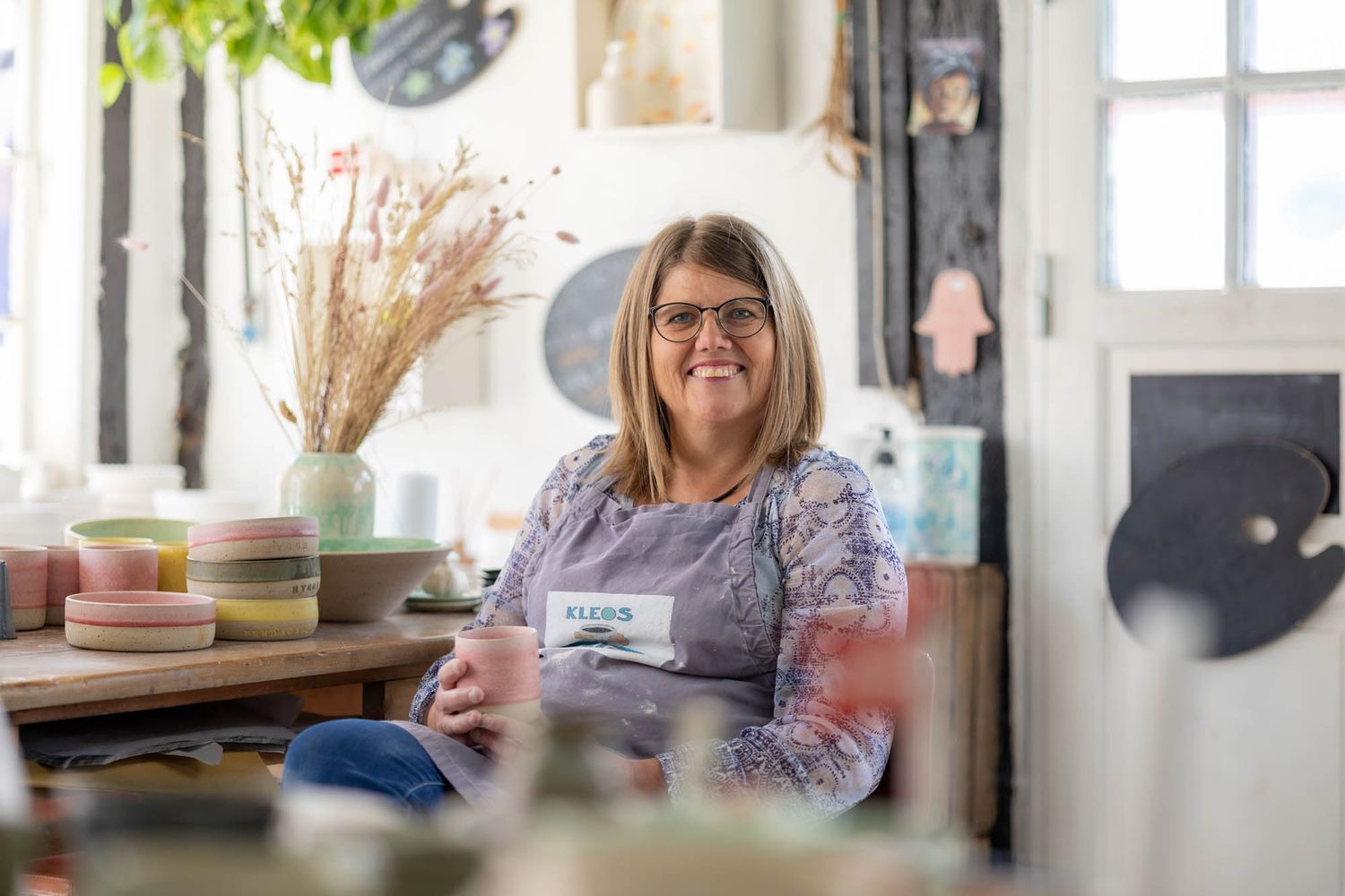 A bright pottery studio workspace with dried flowers and shelving visible in the background.