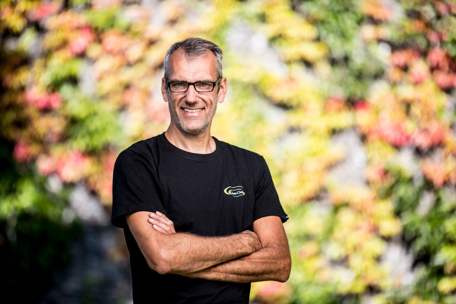 Professional in black shirt standing confidently against colorful autumn foliage.
