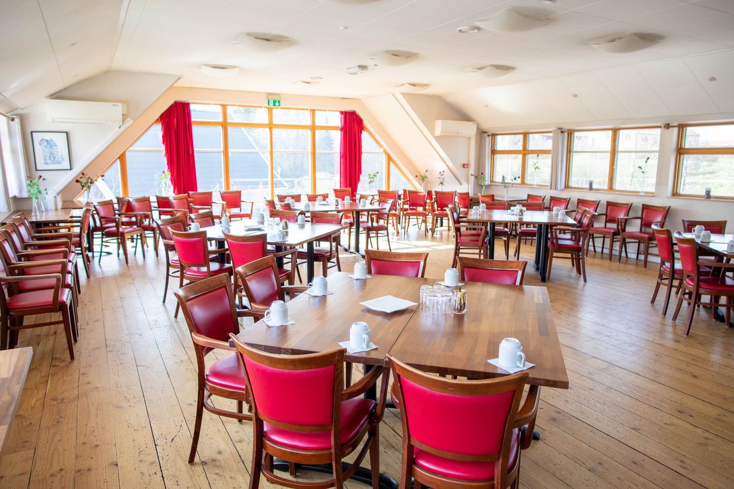 Large bright restaurant interior with red chairs and wooden floors overlooking windows.